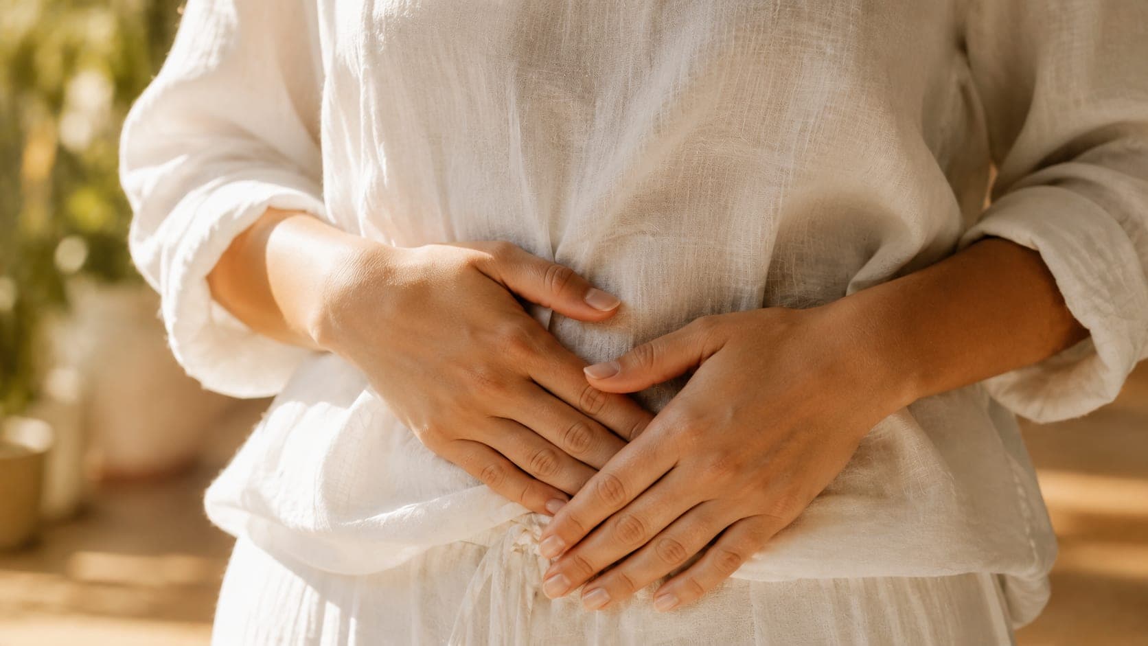 Close-up of a person's hands resting gently on their midsection, wearing a white linen top, soft golden morning light, representing internal gut biology and the L-cell satiety system