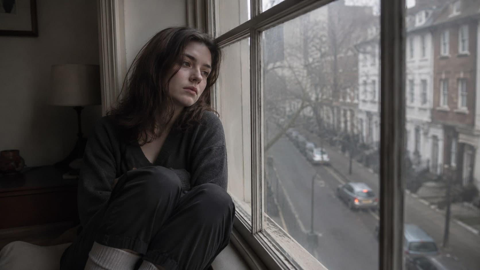 Young woman sitting on a window ledge looking out at a grey rainy street below, wearing dark clothes, arms wrapped around knees, flat and absent expression, muted grey and beige interior tones, still and melancholic atmosphere