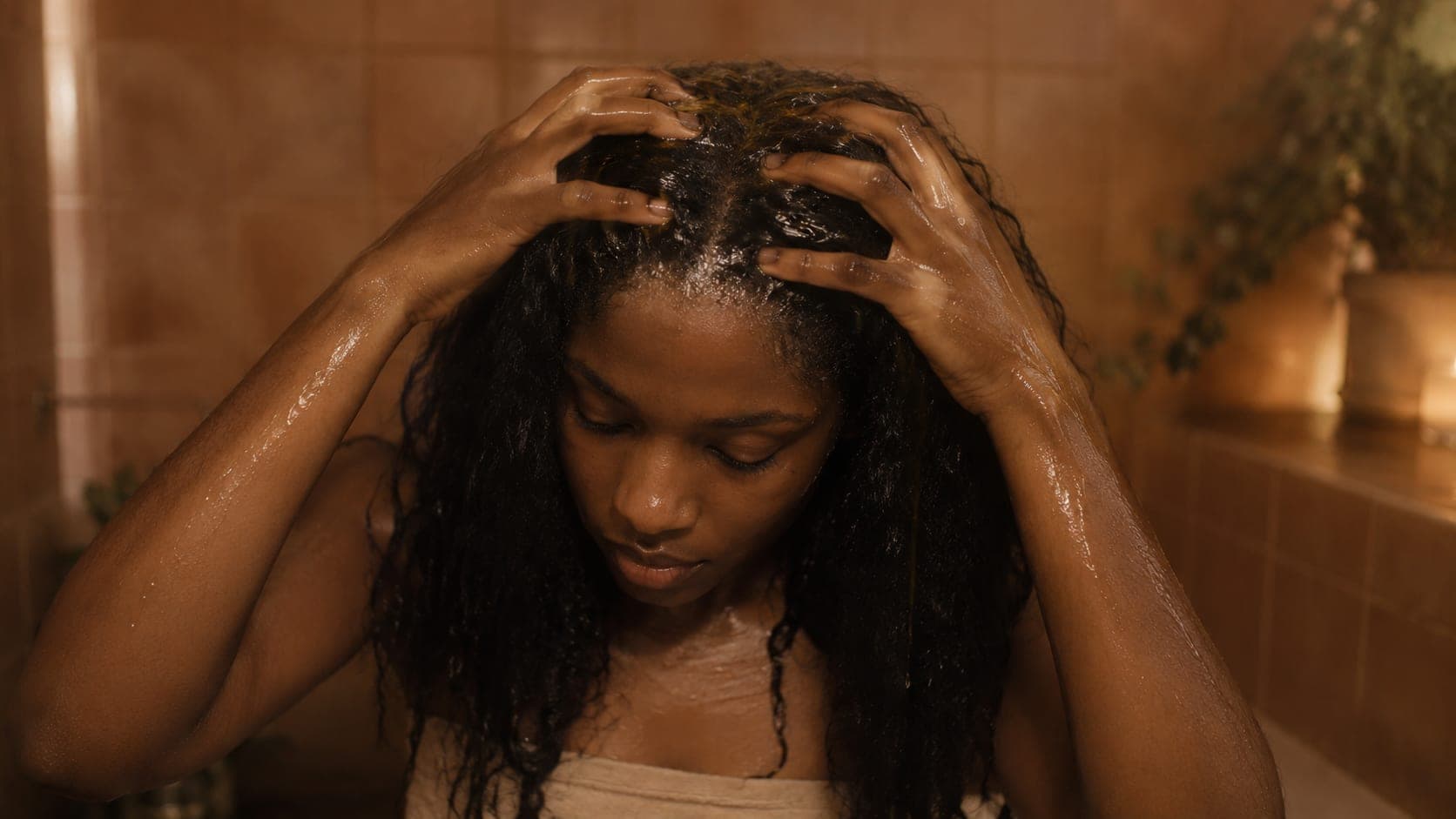 Young woman with dark curly hair applying herbal rinse to her scalp with both hands, circular fingertip movements at the crown, warm amber bathroom light