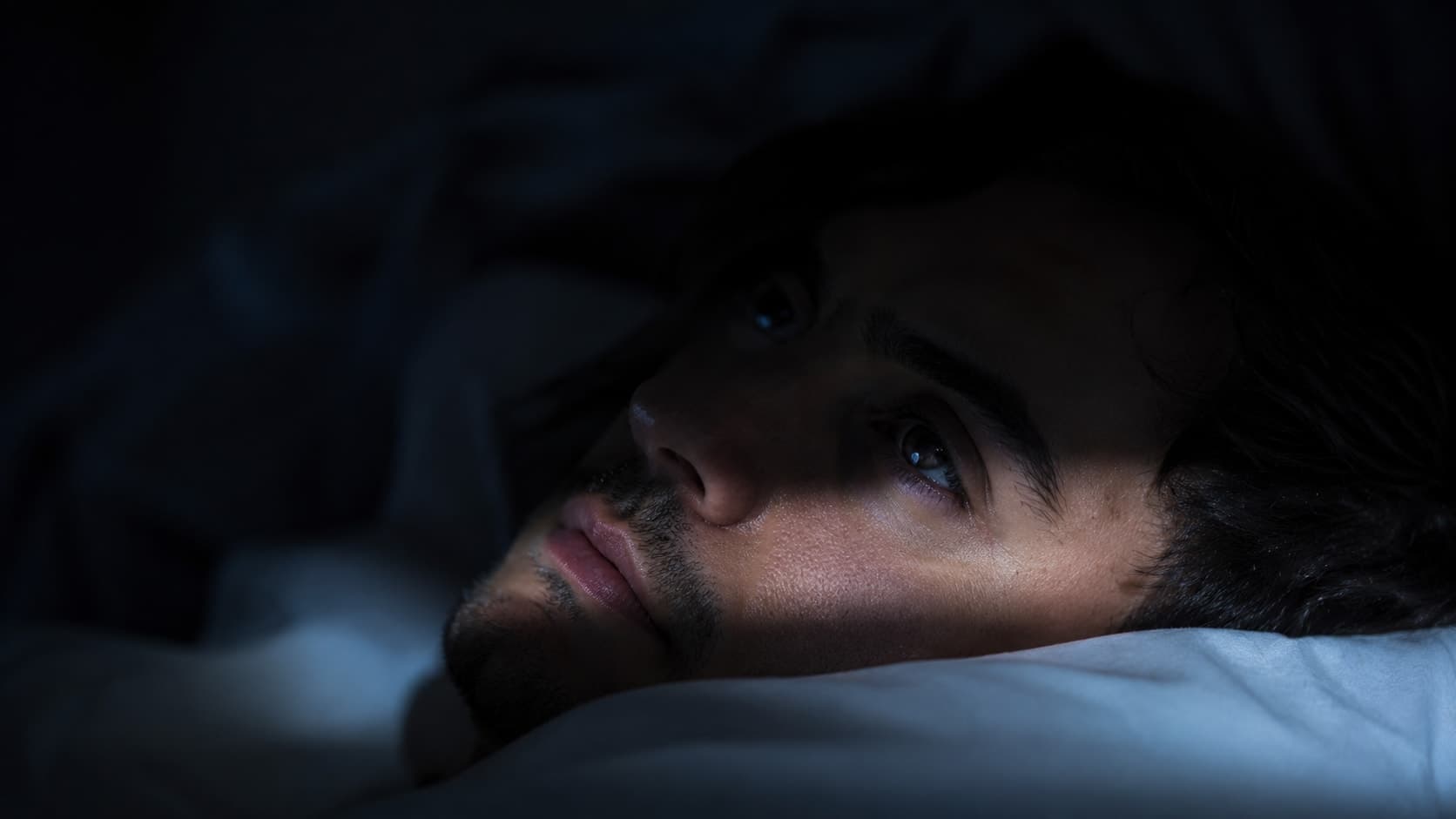 Person lying awake in bed at night with eyes open, lit by cold window light, representing the hyperarousal state that prevents sleep onset despite physical exhaustion
