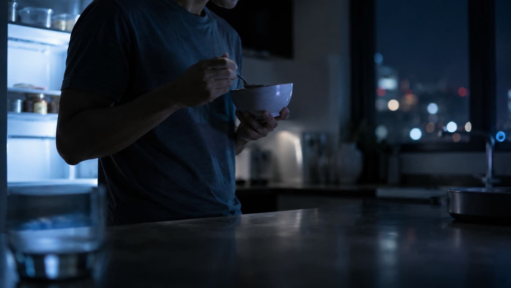 Person eating alone at a dimly lit kitchen counter late at night, illustrating circadian misalignment in eating habits