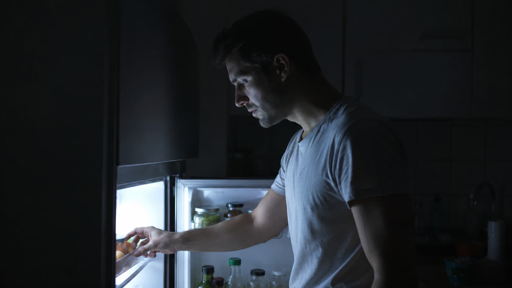 Man in his early 30s at a kitchen counter late at night, refrigerator door open emitting cold blue-white light, reaching for food, cinematic editorial photography, cool blue tones