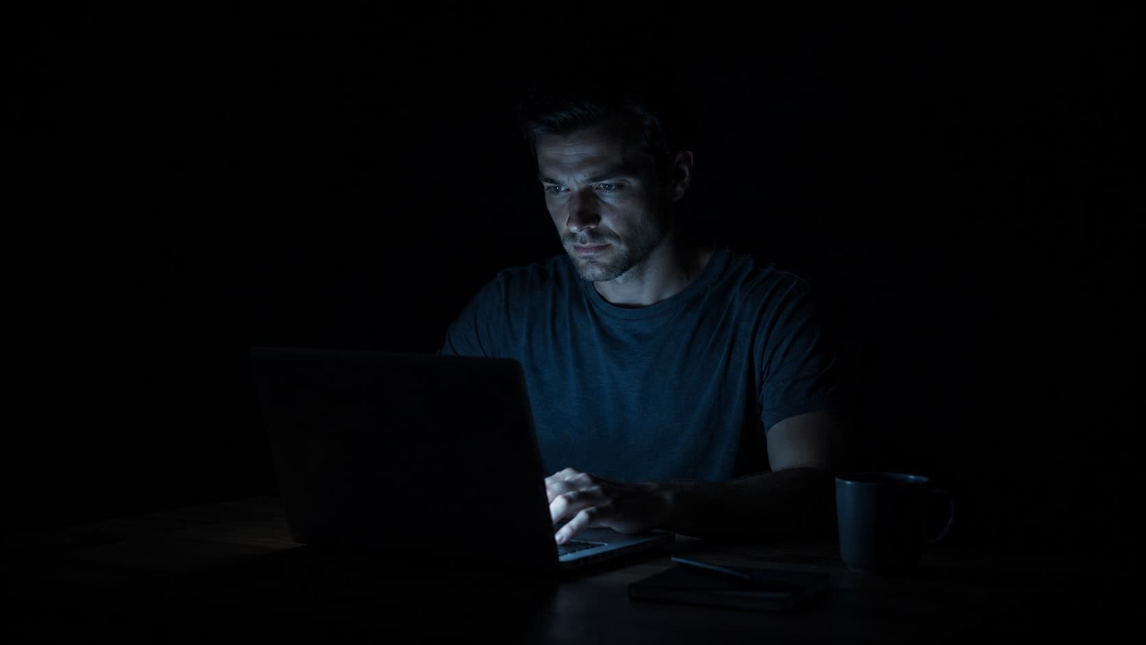 Man in his early 30s at a desk in a dark room, face lit by a bright laptop screen in the evening, representing the evening cortisol elevation sustained by artificial blue light after dark