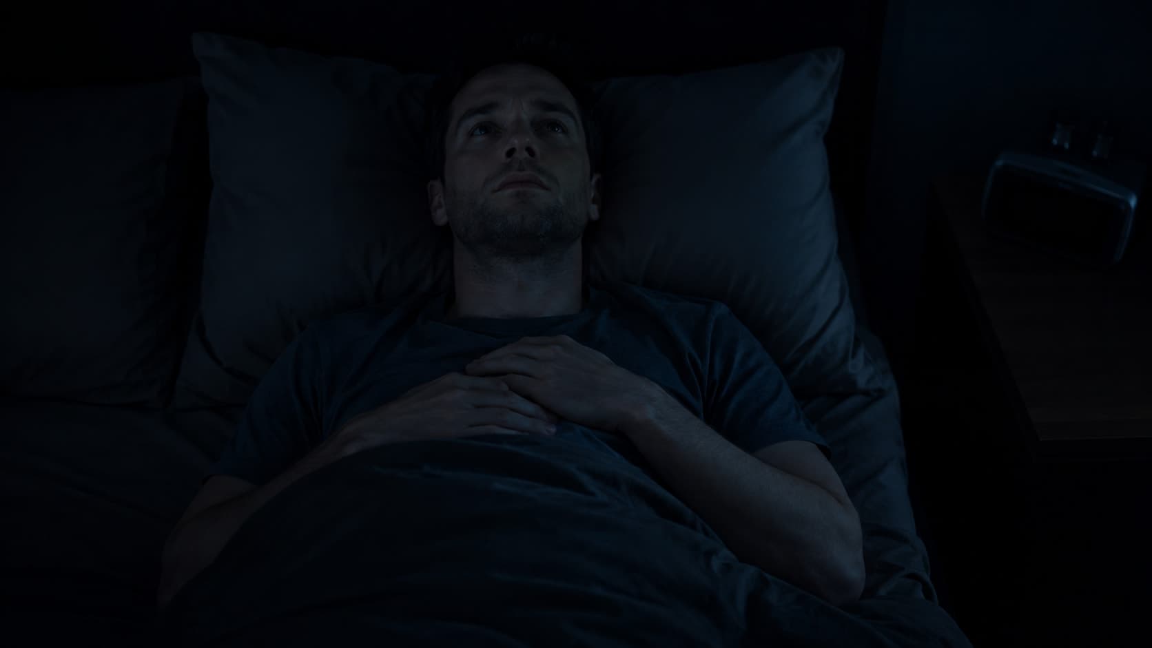 Man in his early 30s sitting on the edge of a bed in darkness, a digital clock on the nightstand showing 3:00 AM, representing the premature cortisol surge that triggers early waking