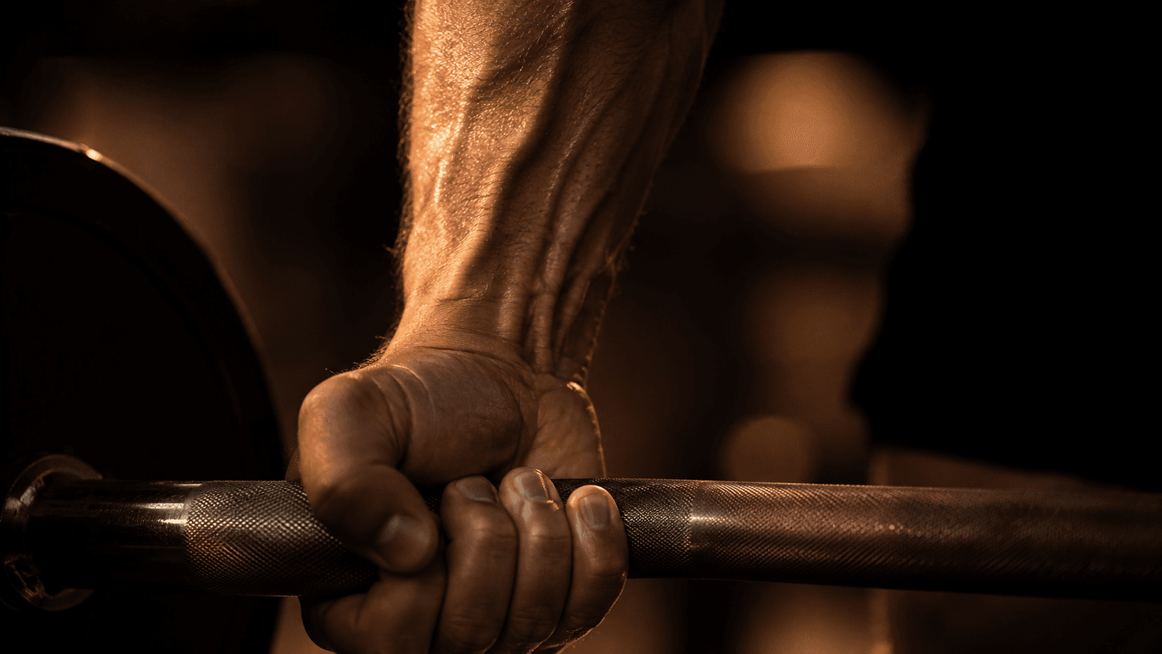 Extreme close-up of a muscular hand and forearm gripping a barbell with warm amber gym lighting, showing muscle definition, veins and texture under dramatic side lighting