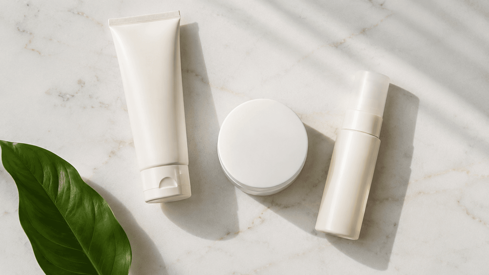 Overhead flat lay of three minimalist white skincare products on white marble with warm morning window light and a fresh green leaf — a cleanser, ceramide moisturizer, and SPF